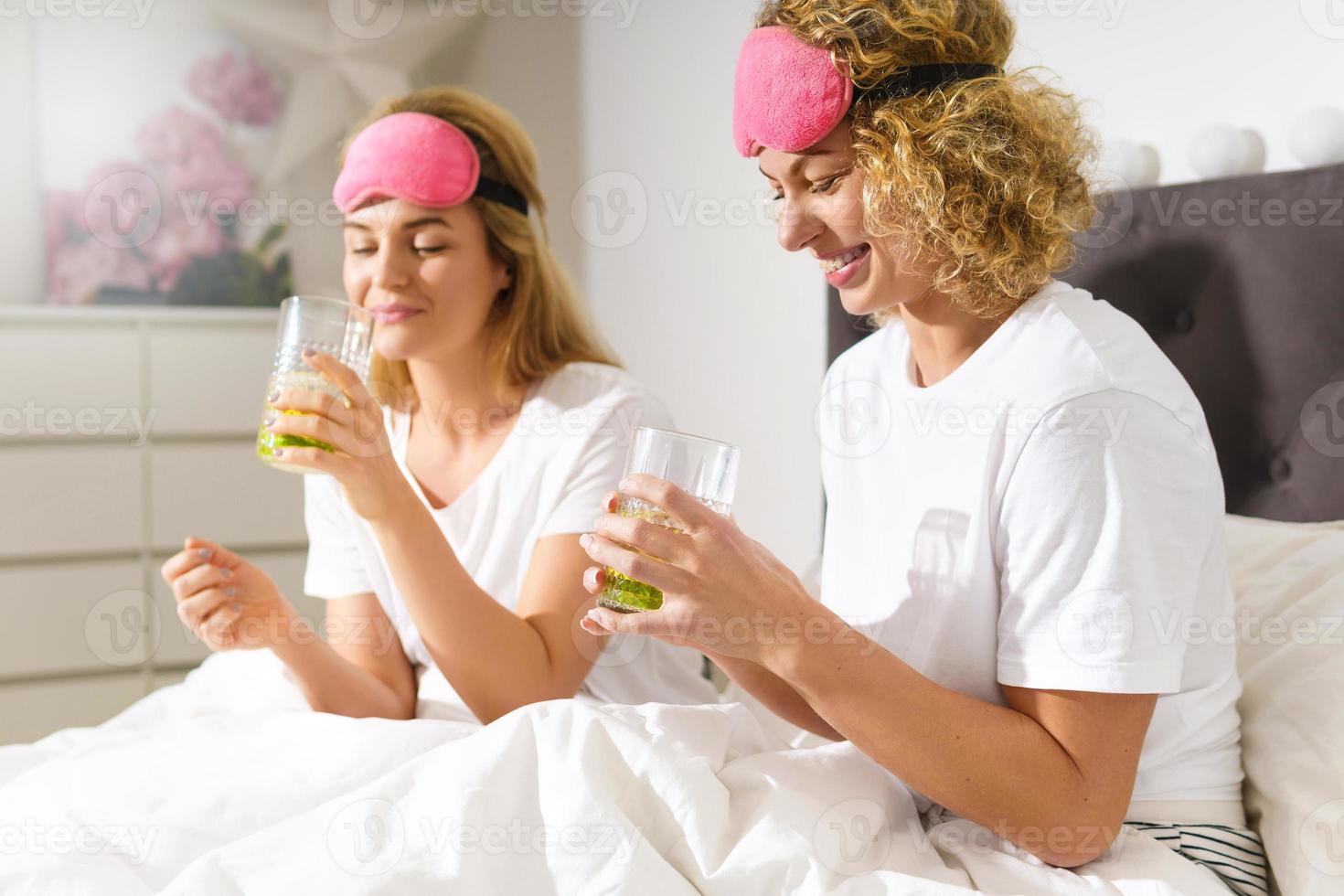 Two beautiful and cheerful women drinking detox water after awakening in comfortable bed photo