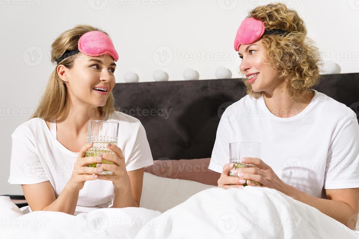 Two beautiful and cheerful women drinking detox water after awakening in comfortable bed photo