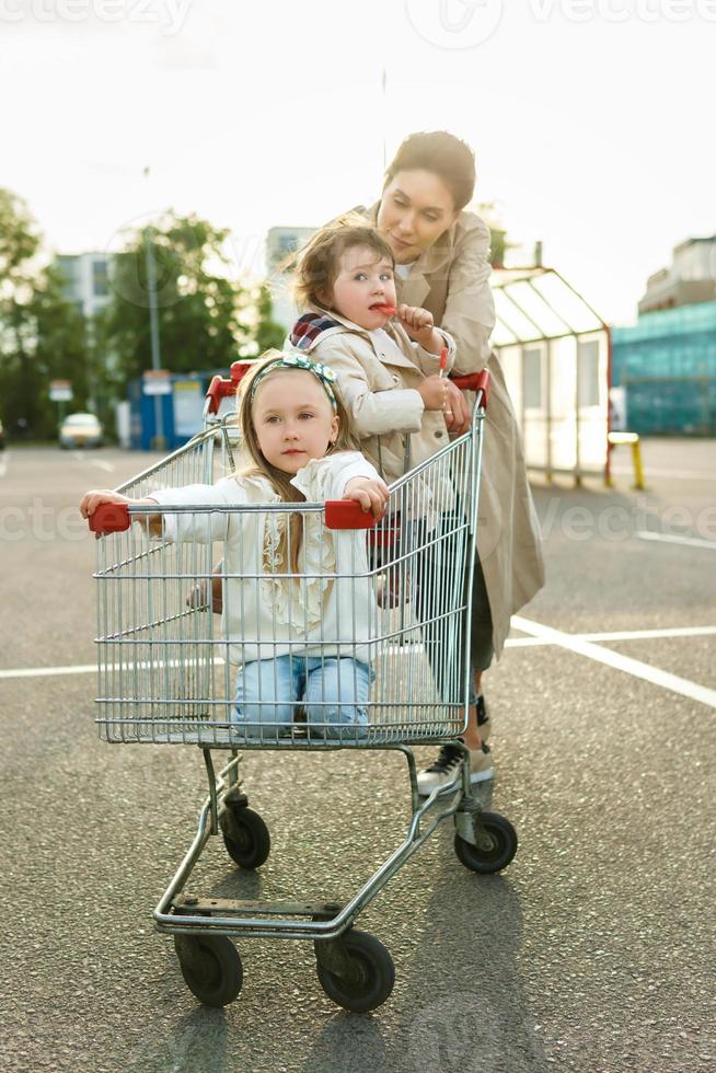 feliz madre y sus hijas se divierten con un carrito de compras en un estacionamiento al lado de un supermercado. foto