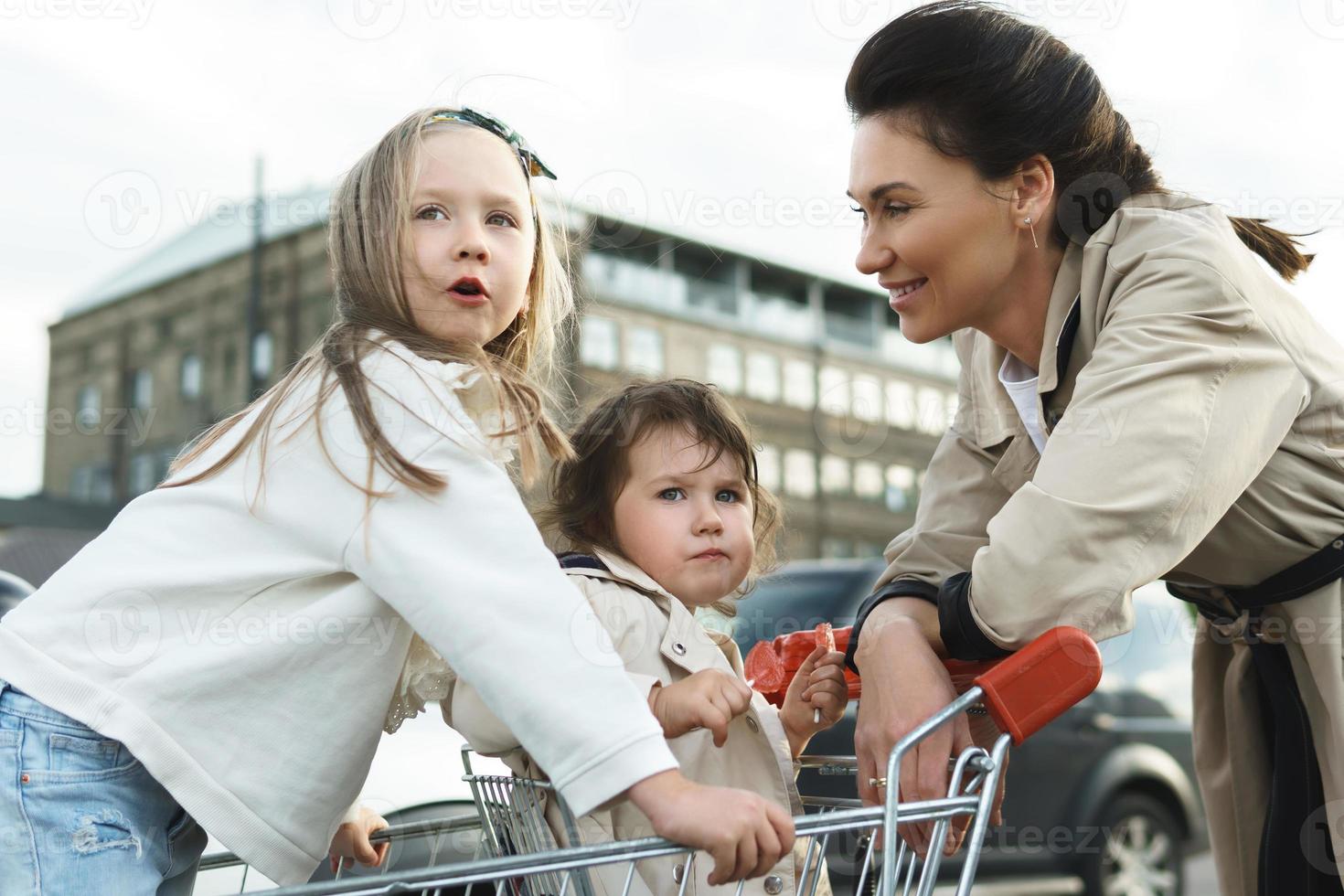 feliz madre y sus hijas se divierten con un carrito de compras en un estacionamiento al lado de un supermercado. foto