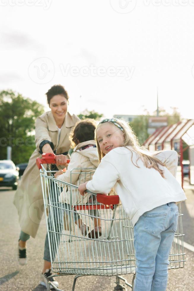 Happy mother and her daughters are having fun with a shopping cart on a parking lot beside a supermarket. photo