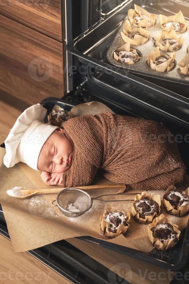 Newborn baby wearing chef's hat is lying on the oven tray with a muffins photo