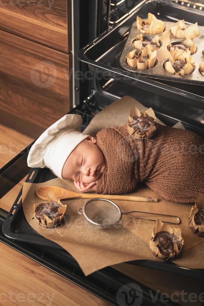 un bebé recién nacido con sombrero de chef está tirado en la bandeja del horno con muffins foto