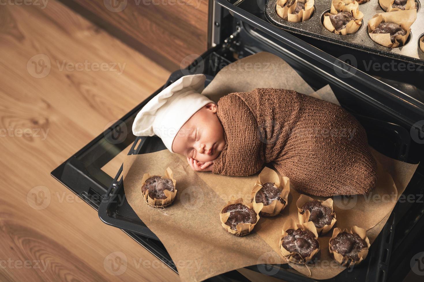 Newborn baby wearing chef's hat is lying on the oven tray with a muffins photo