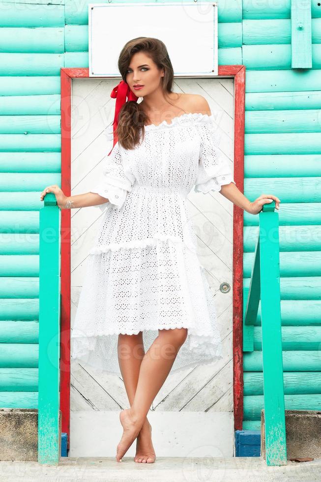 Cute woman wearing beautiful white dress is posing near colorful wooden house photo