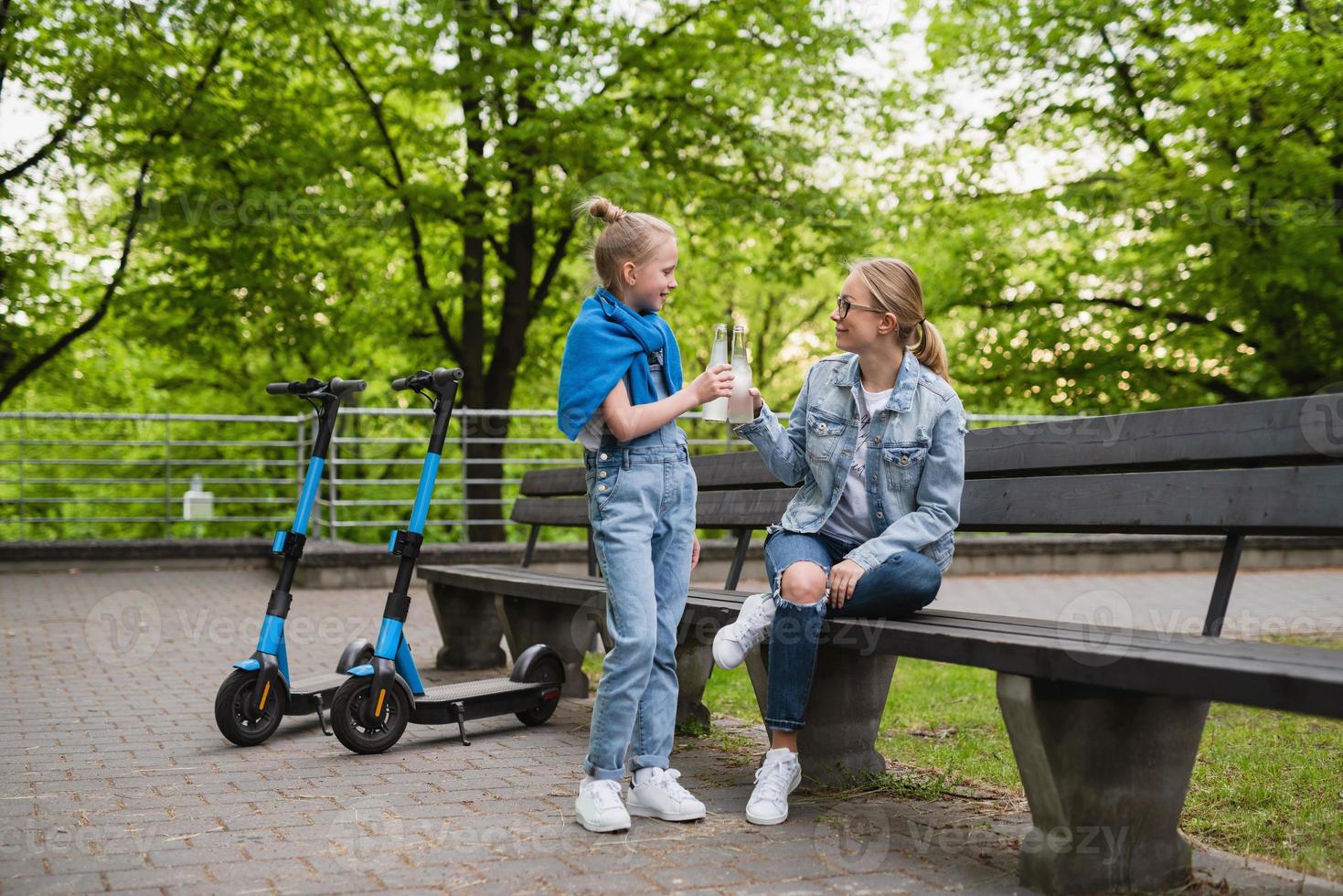 Mother and daughter with electric scooters are drinking lemonade in city park photo
