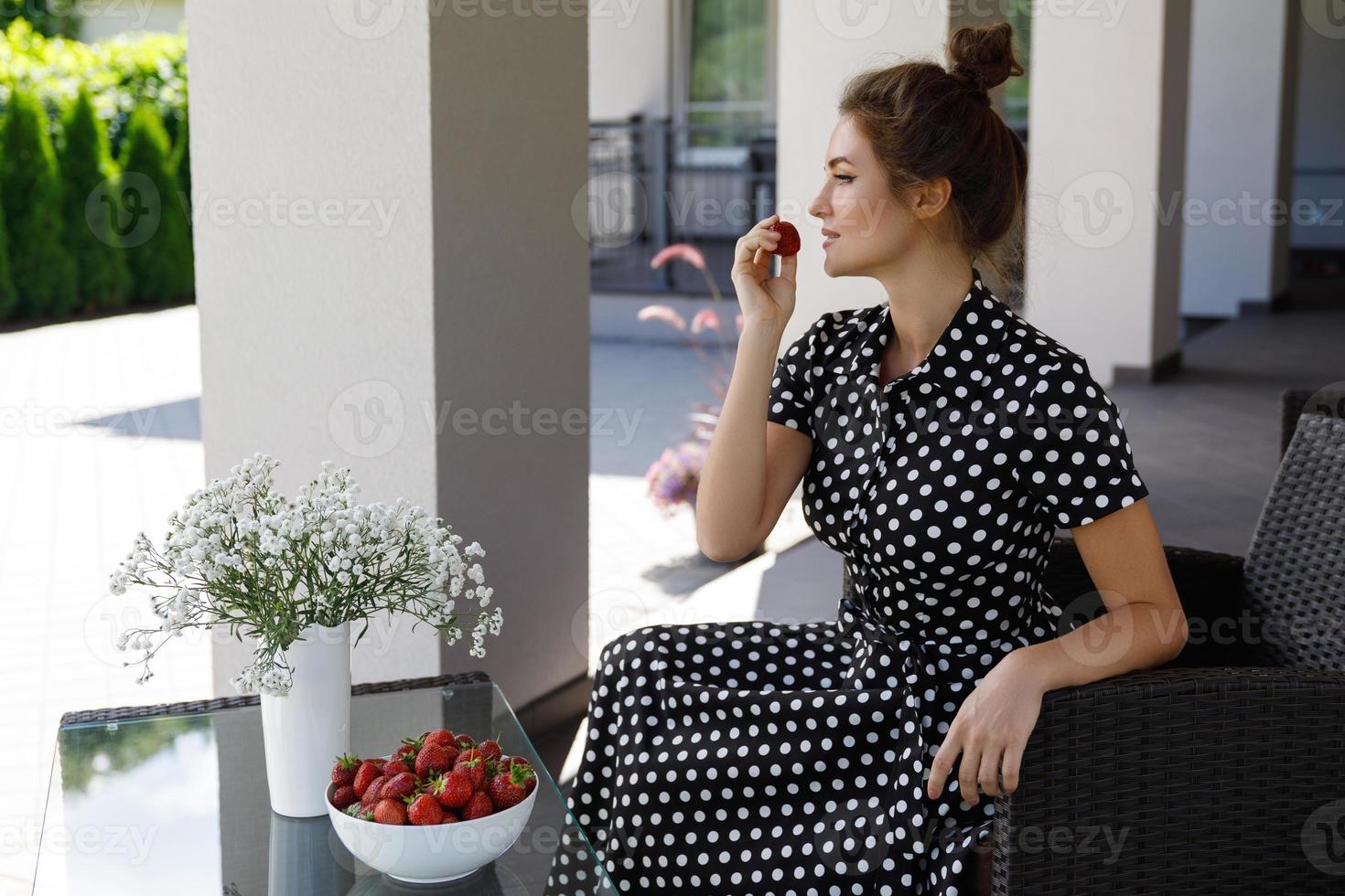 Gorgeous woman wearing beautiful dress with a polka dot pattern sitting in a patio and eating strawberry photo