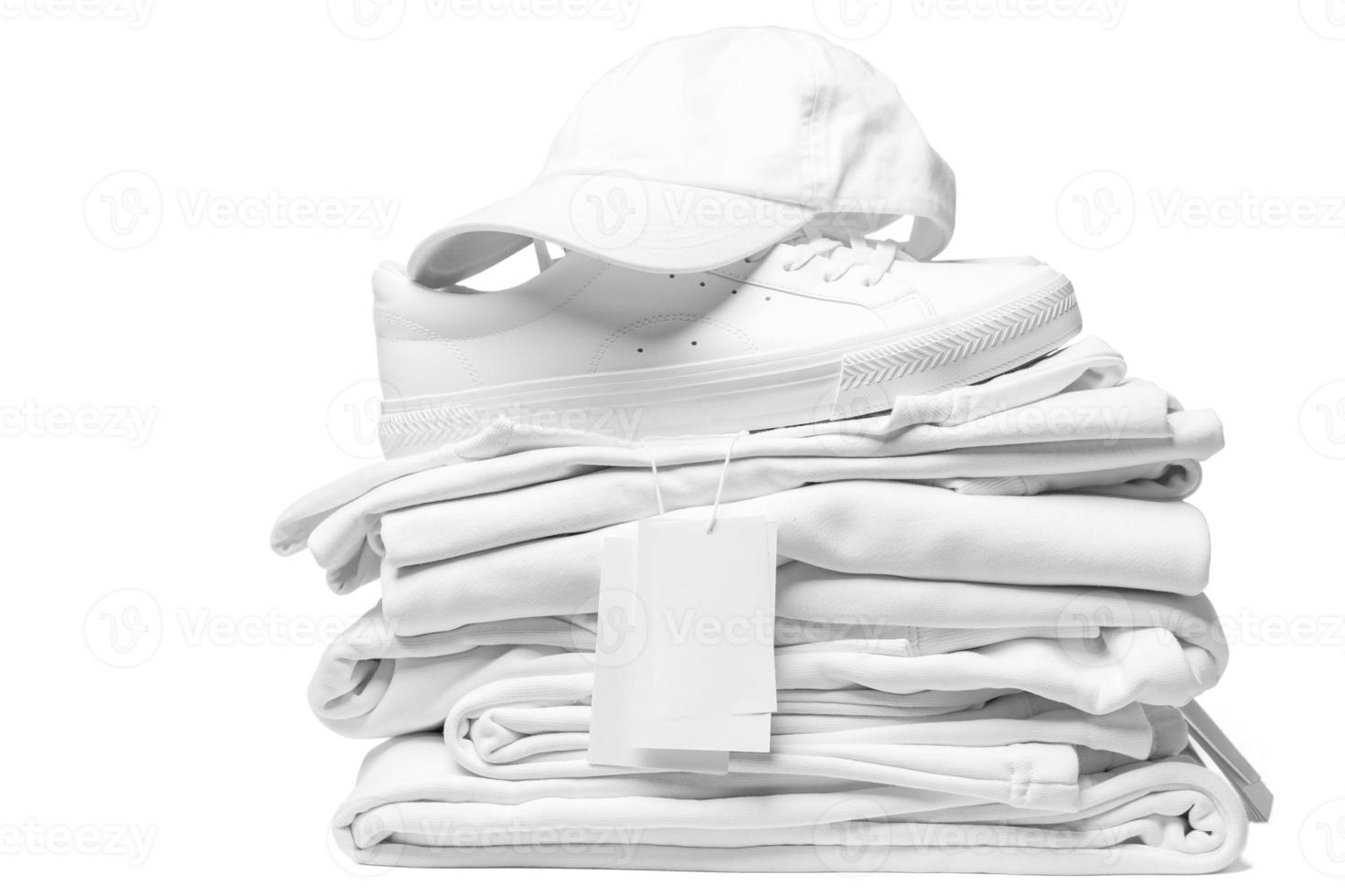 Stack of white clothes, trainers and baseball cap with a blank garment tag on white background photo