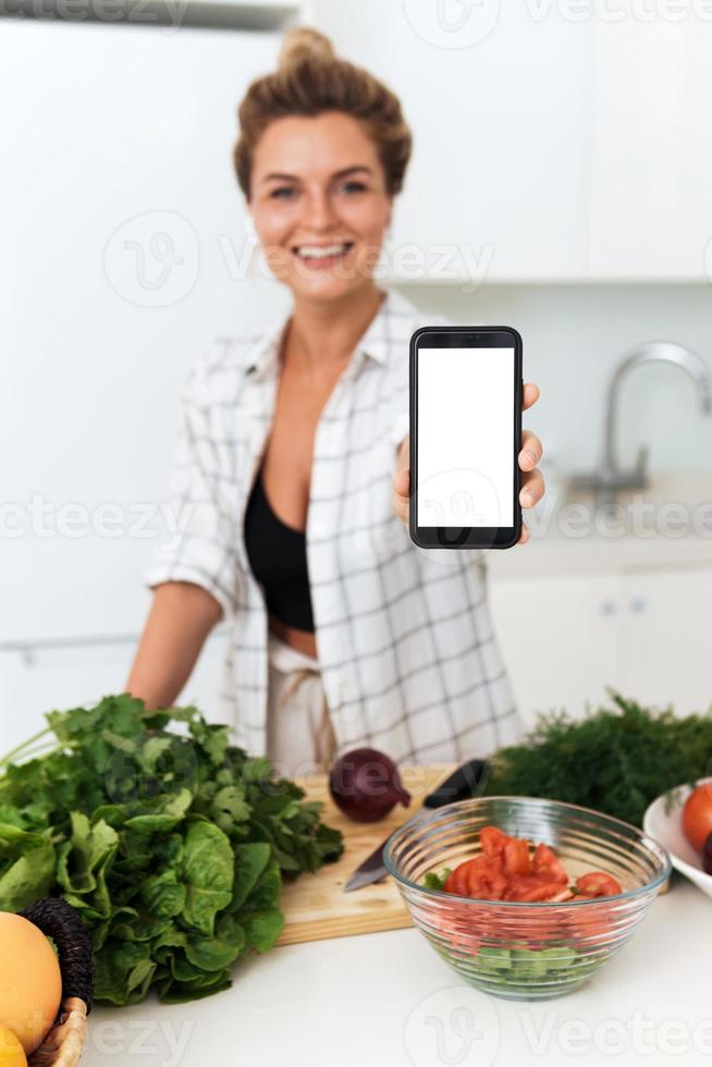 Woman is holding smartphone with blank screen for your design during cooking in modern kitchen photo