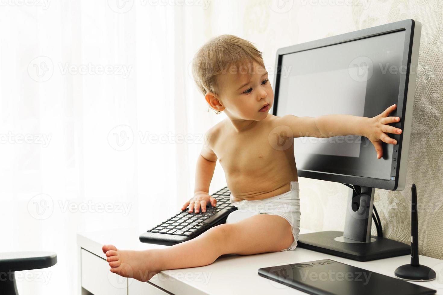 Little boy sitting on a desk and playing with a keyboard and monitor. photo