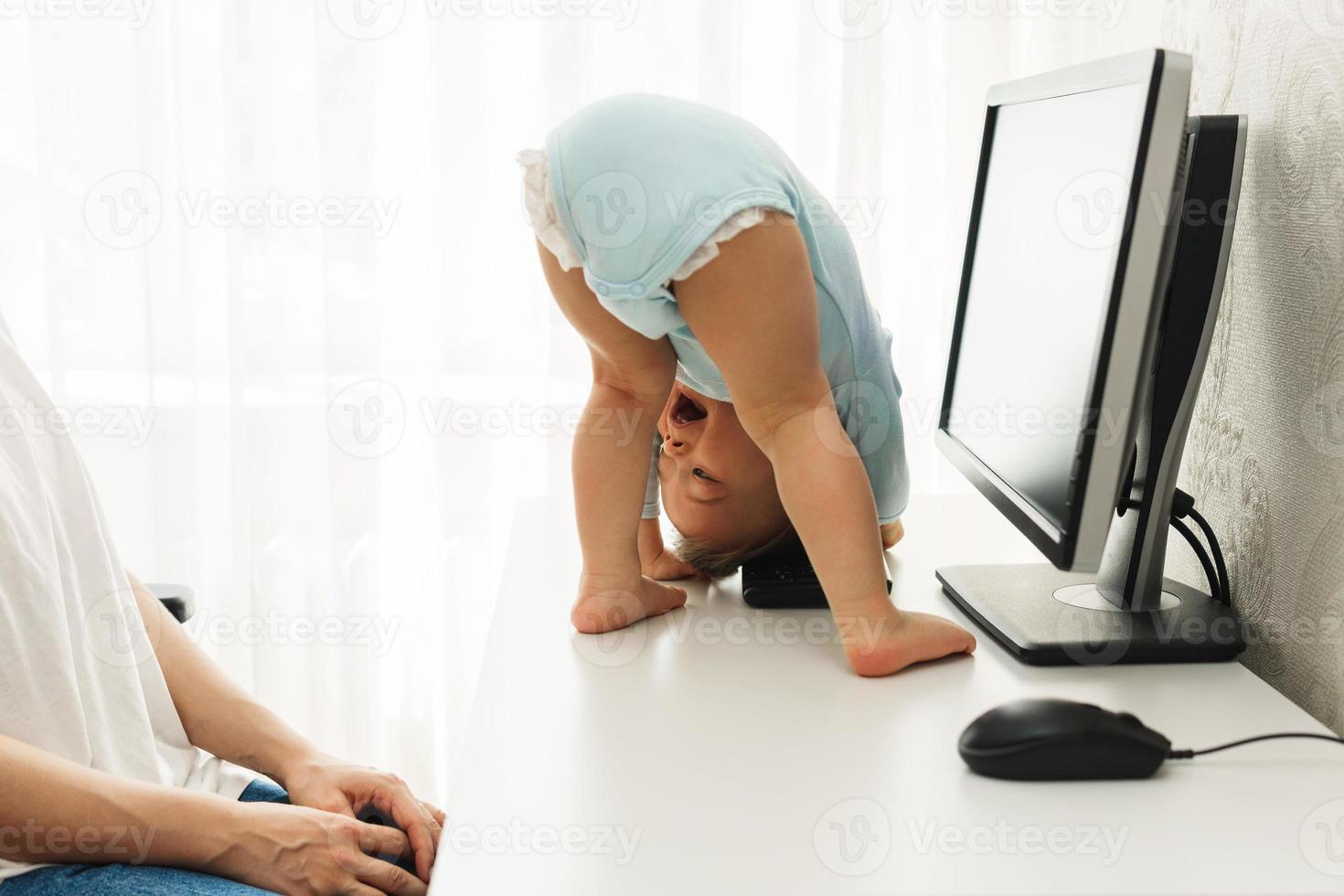 Little boy standing upside down on a desk and distracting mother from work on computer. photo