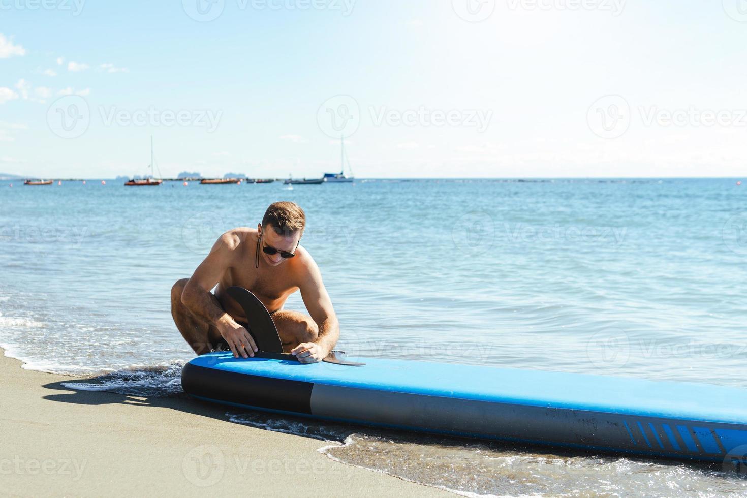 Young male surfer setting up standup paddleboard on a beach. photo
