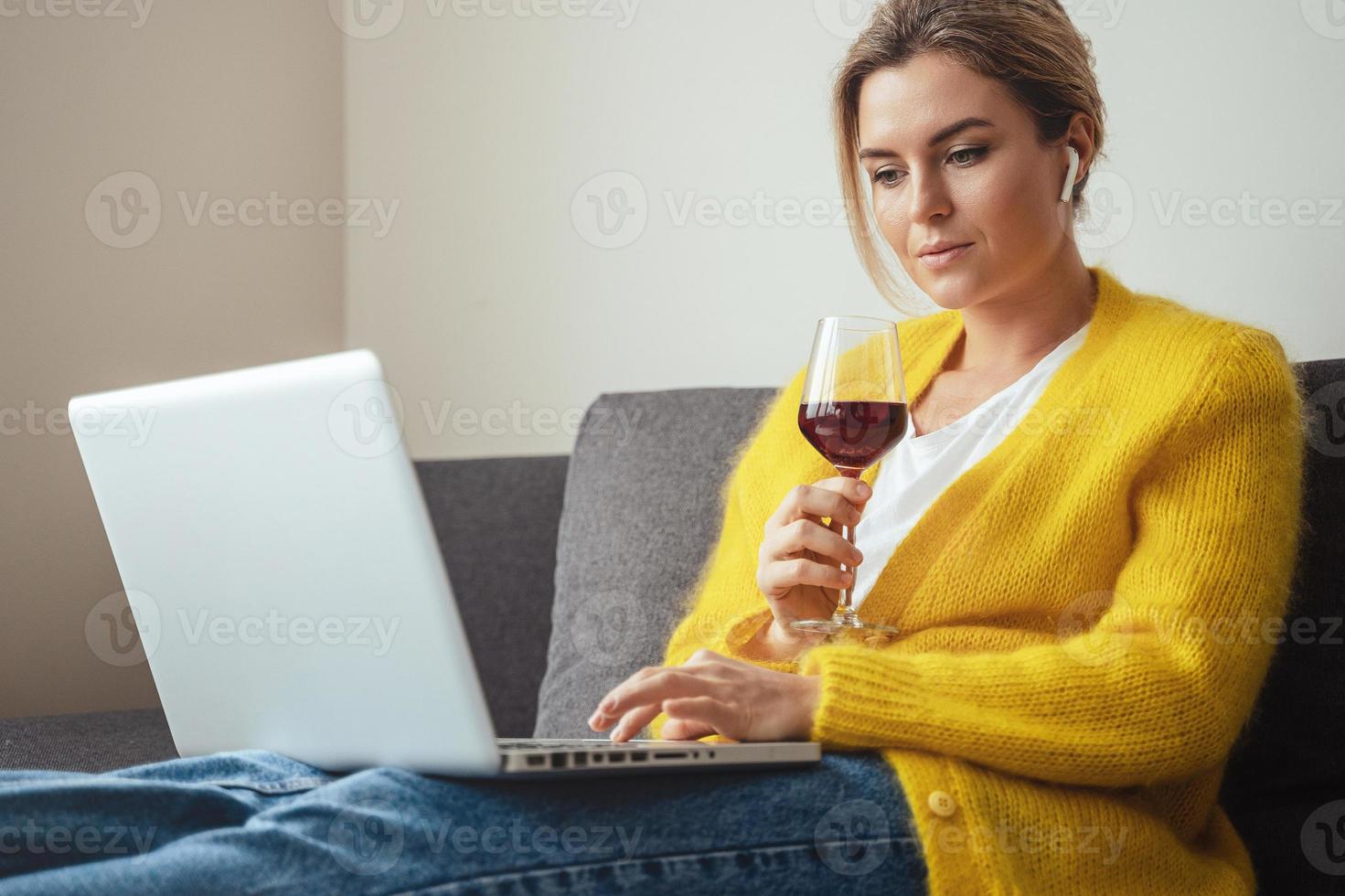 Woman sitting on the sofa drinking red whine while watching movie or working on her laptop at home photo