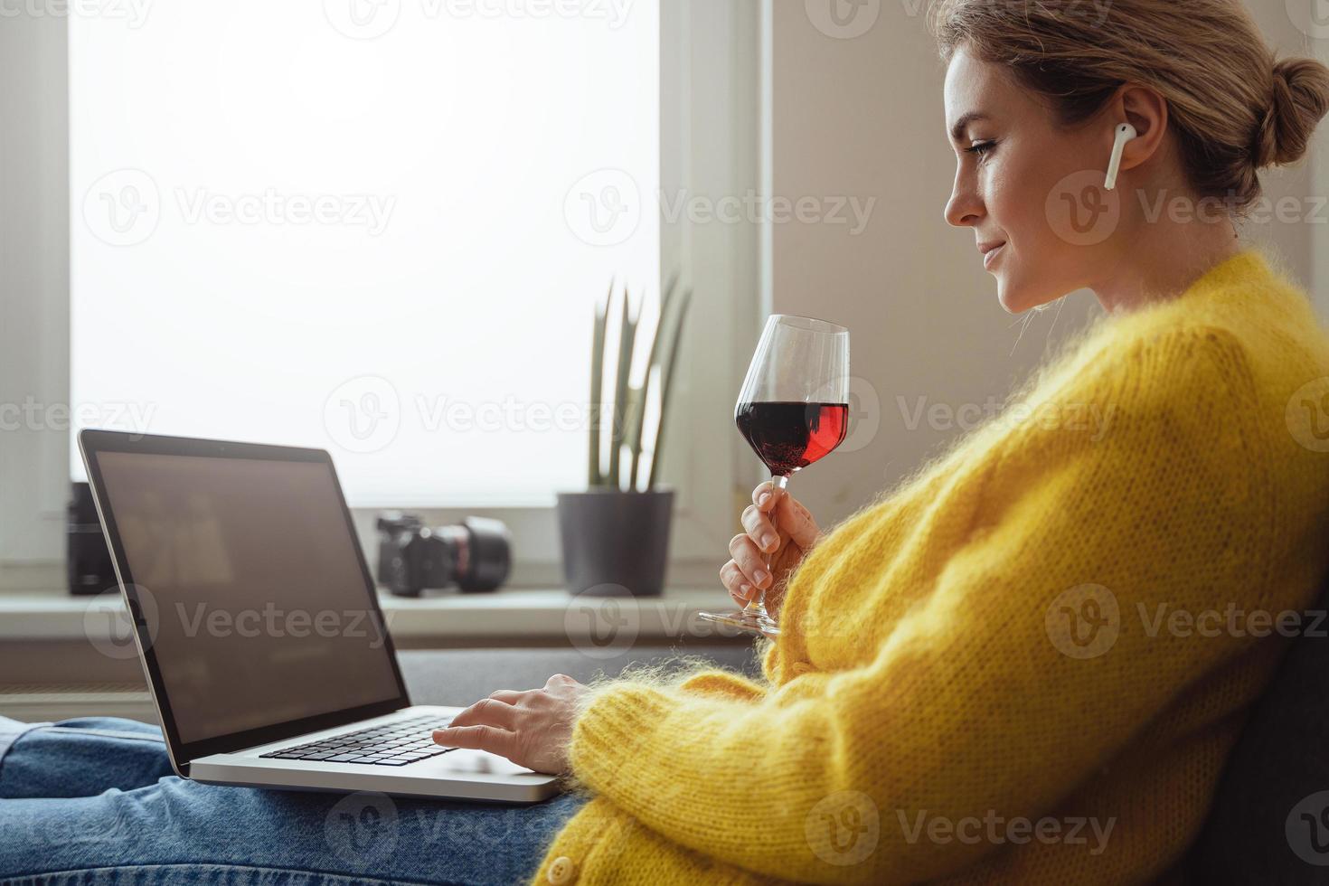 Woman sitting on the sofa drinking red whine while watching movie or working on her laptop at home photo