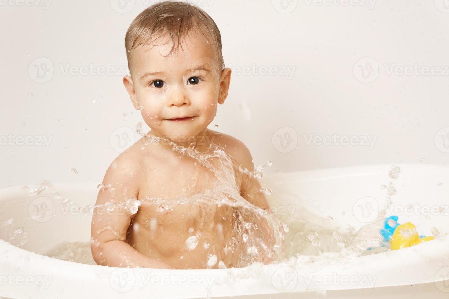 niño pequeño salpicando agua en un baño con juguetes de goma. foto