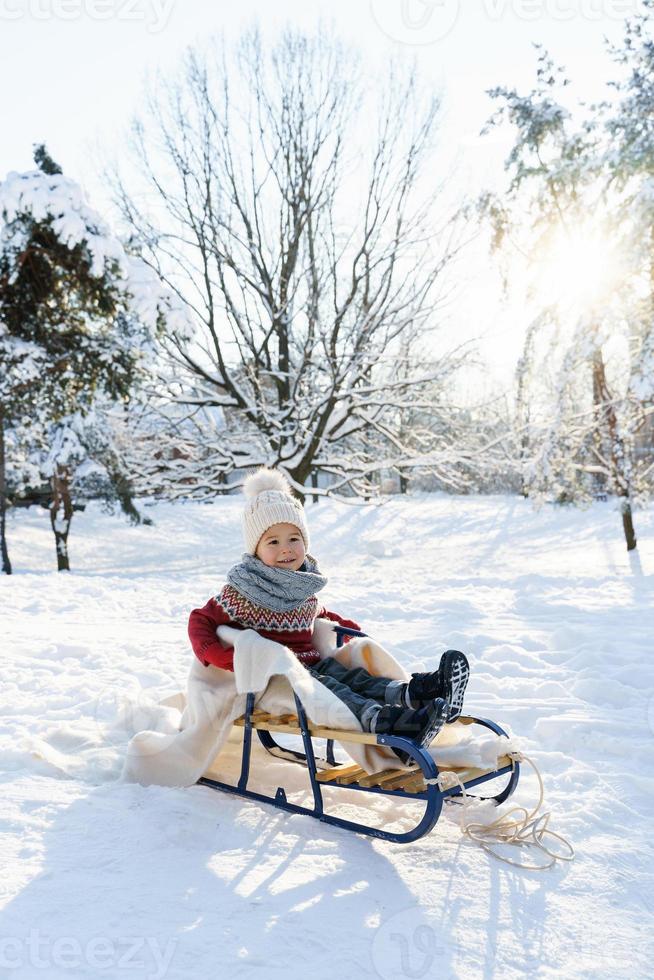 Toddler boy sitting on the sleigh in a snowy city park during sunny winter day photo