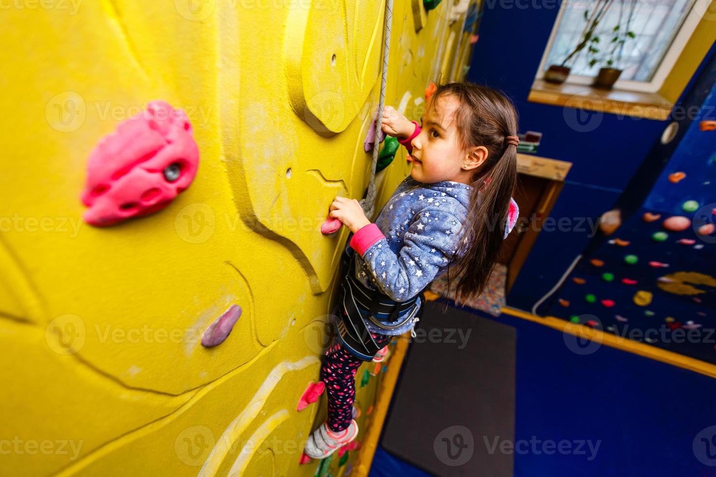 Little baby girl with funny hear style climbing vertical wall and man belaying her from below photo