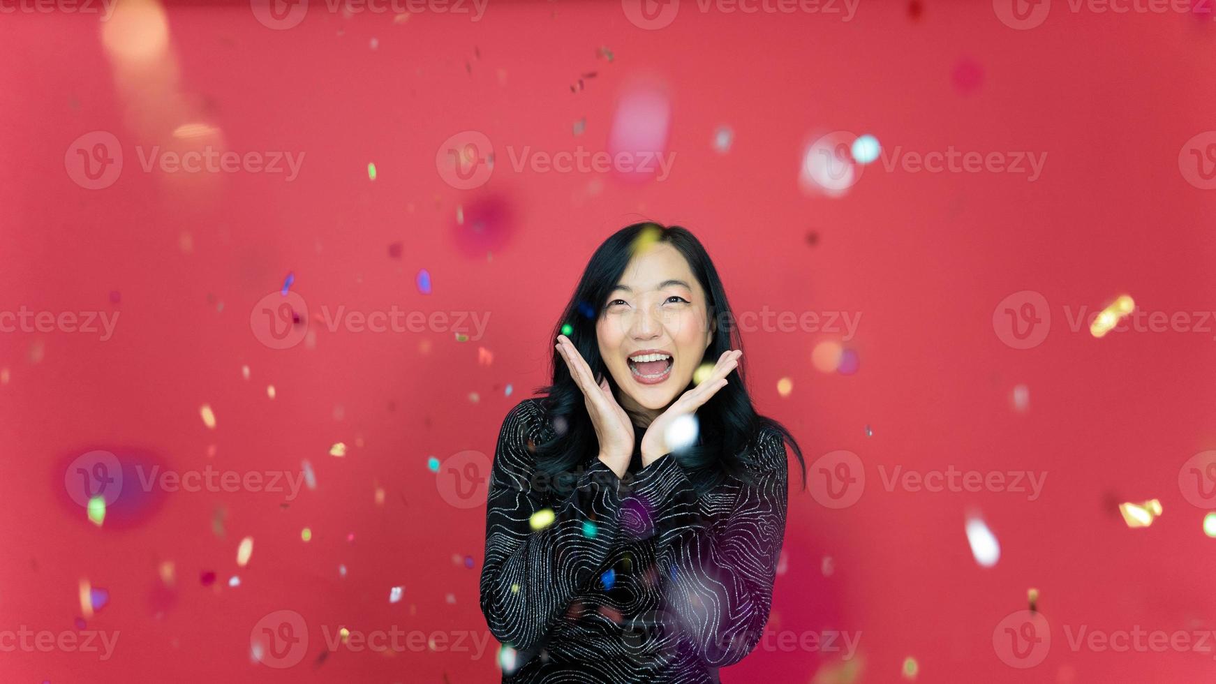 Holiday, new year and party concept. Beautiful young woman wearing evening dress standing under confetti rain over red background. Happy time. photo