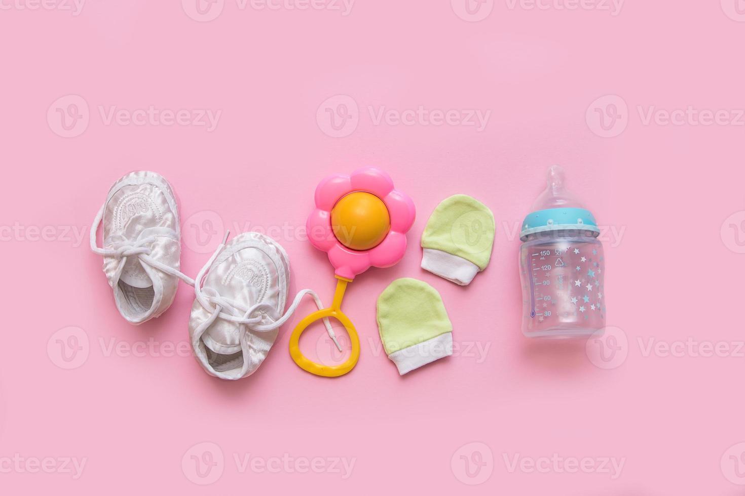 accesorios para un recién nacido - botines, un sonajero, mitones y una botella de fórmula sobre un fondo rosa foto