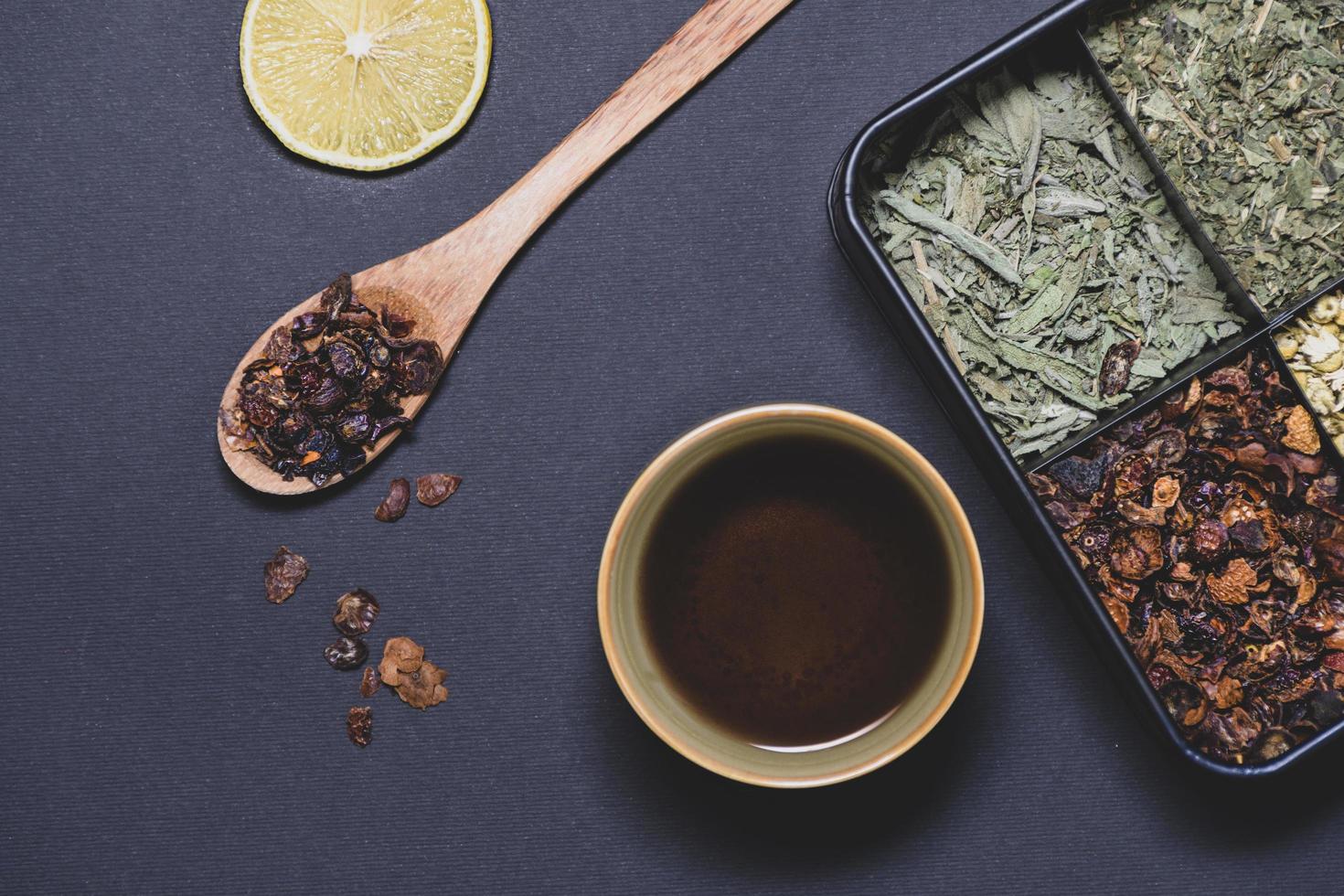 A cup of tea next to a wooden spoon, a slice of lemon and a box full of different tea leaves photo