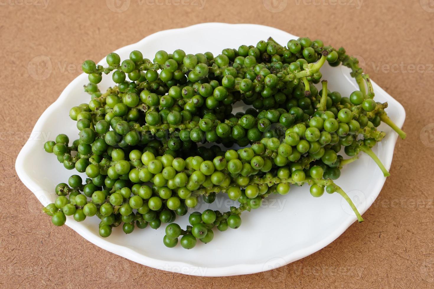 Green young Thai pepper fruits on white plate. Brown wooden background. Concept, herbal spices for cooking , food ingredient can be cooked in verious Thai food menu. photo