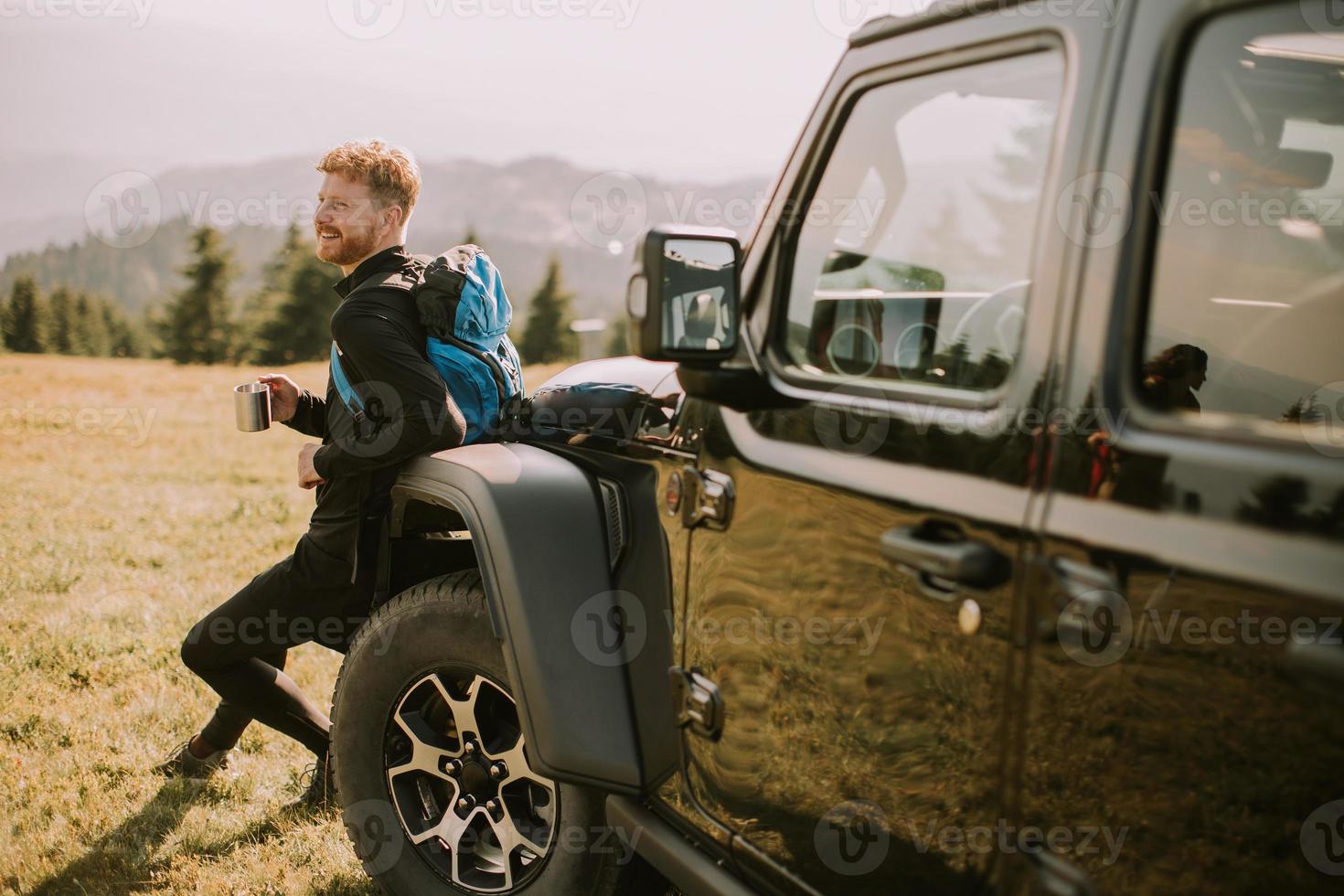 Young man relaxing and drinking coffee by the terrain vehicle hood at countryside photo