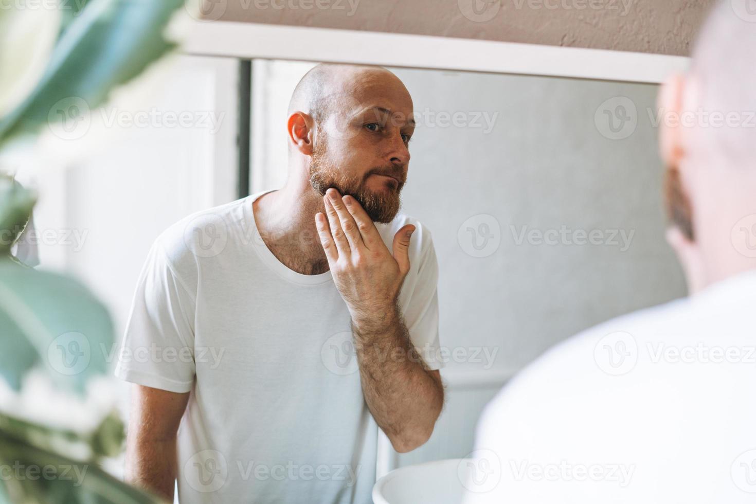Handsome bald man looking at mirror and touching face in bathroom photo