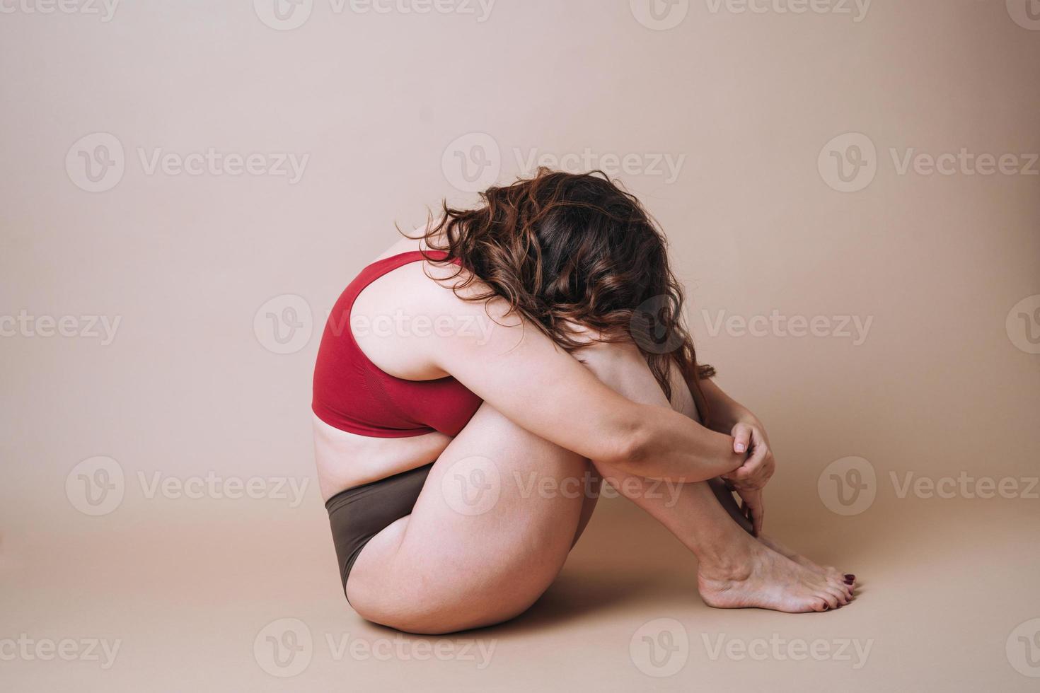 Young unhappy woman plus size in underwear sitting in closed pose on beige background photo