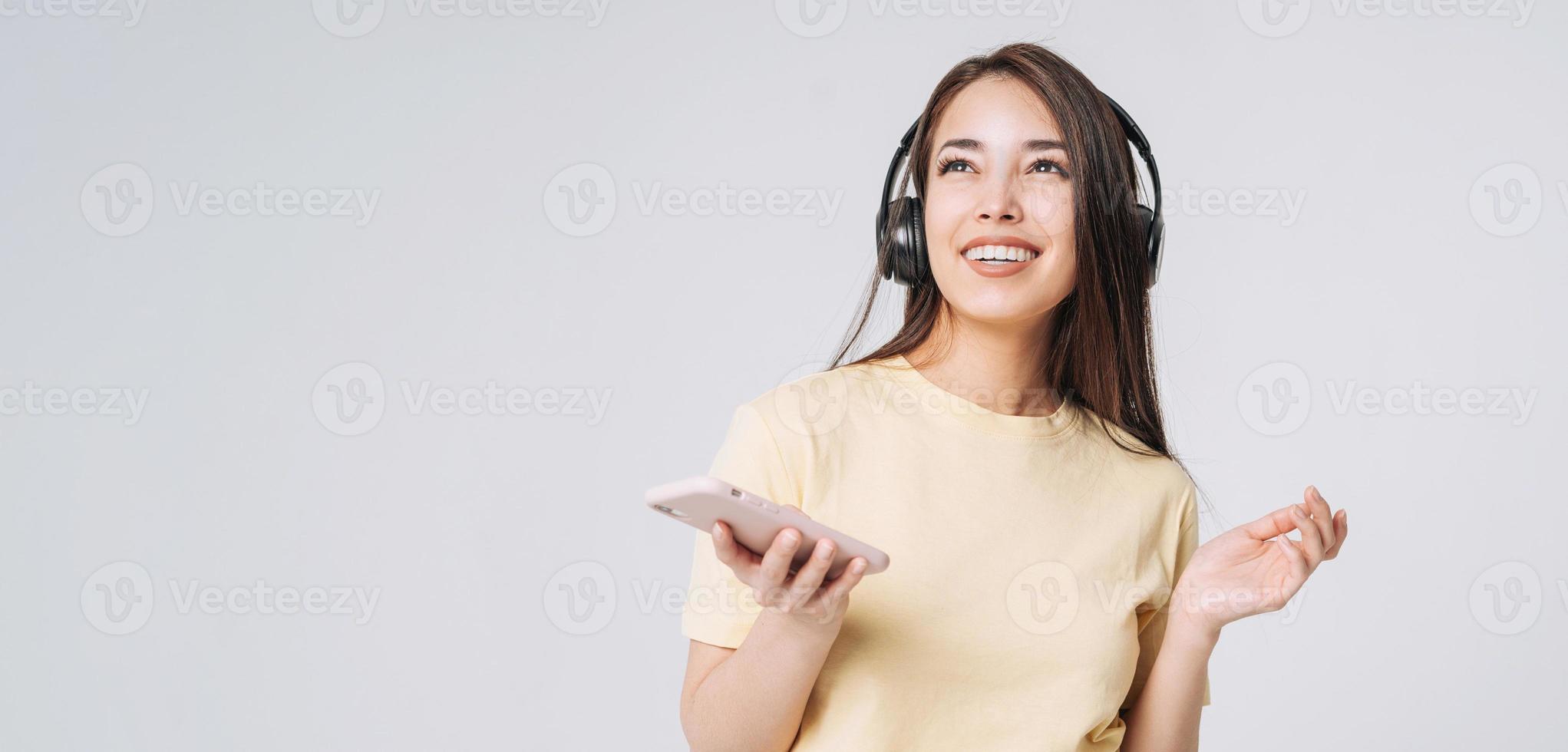 Young happy asian woman with long hair in yellow shirt and jeans using mobile phone listening music by headphones on grey background, banner photo