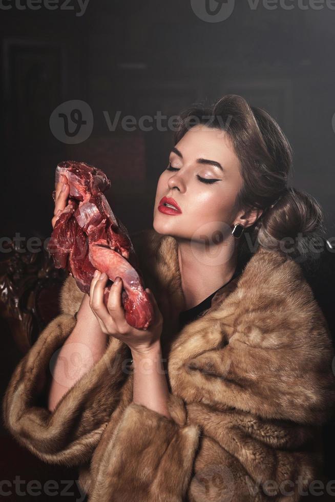 Provocative image of gorgeous wearing fur coat and holding piece of raw beef. Animal rights concept. photo