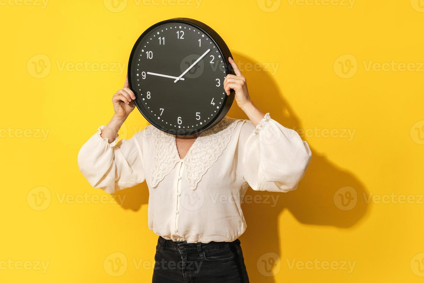 Woman covering her face with big clock on yellow background. photo