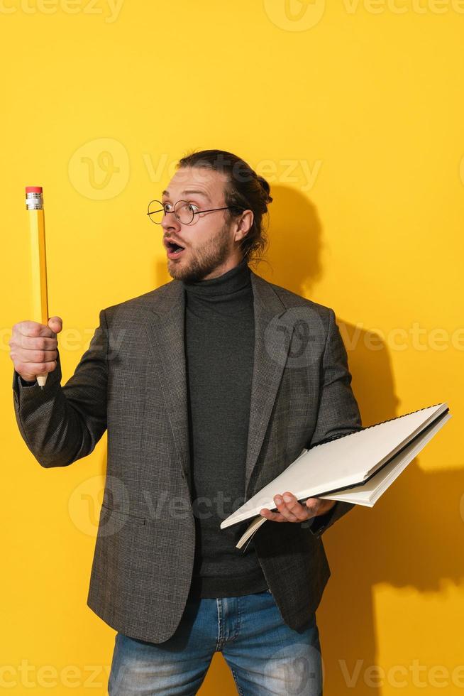 Funny man wearing glasses holding big pencil and notebook on yellow background photo