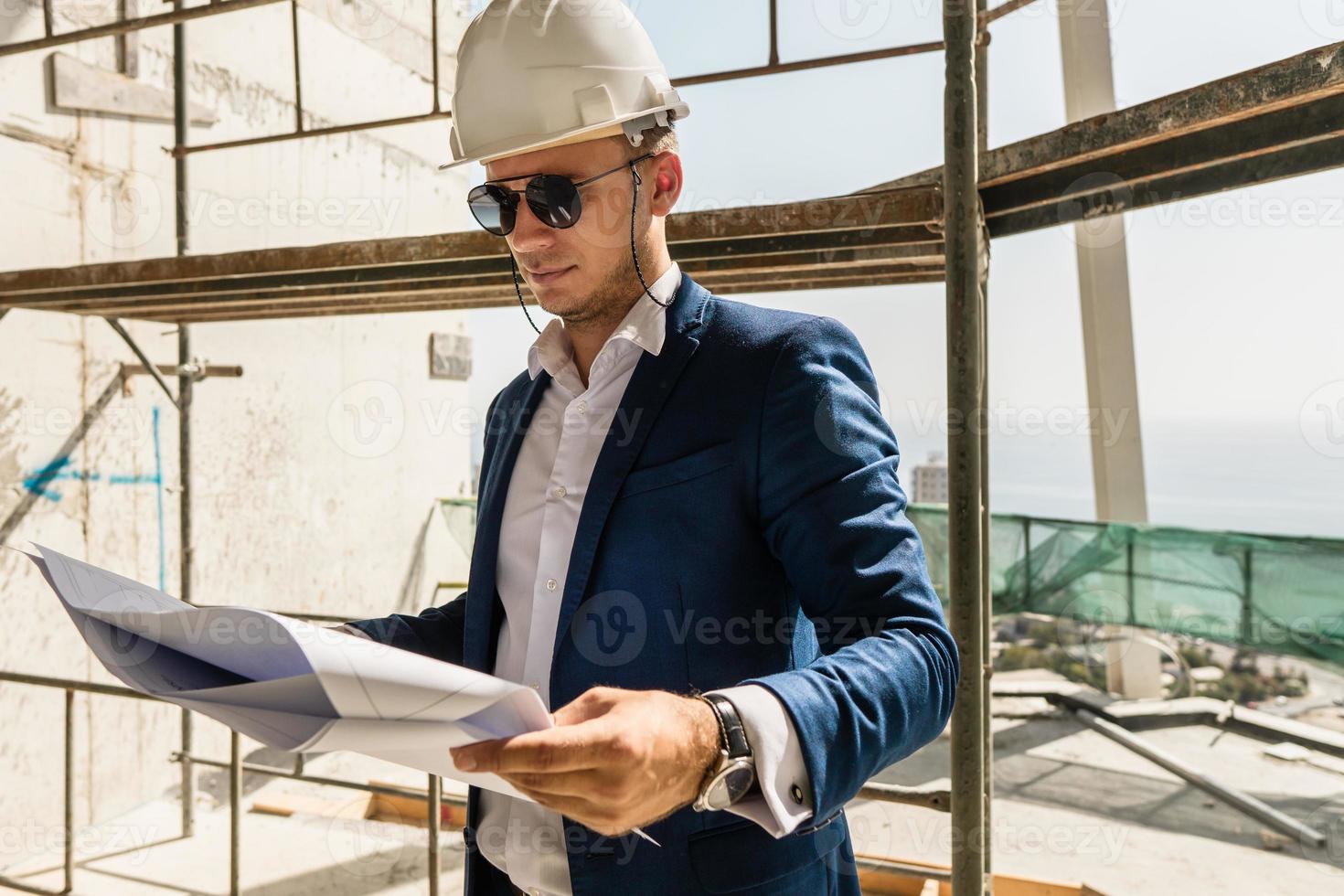 Architect wearing formal suit and hard hatholding a blueprints on a construction site photo