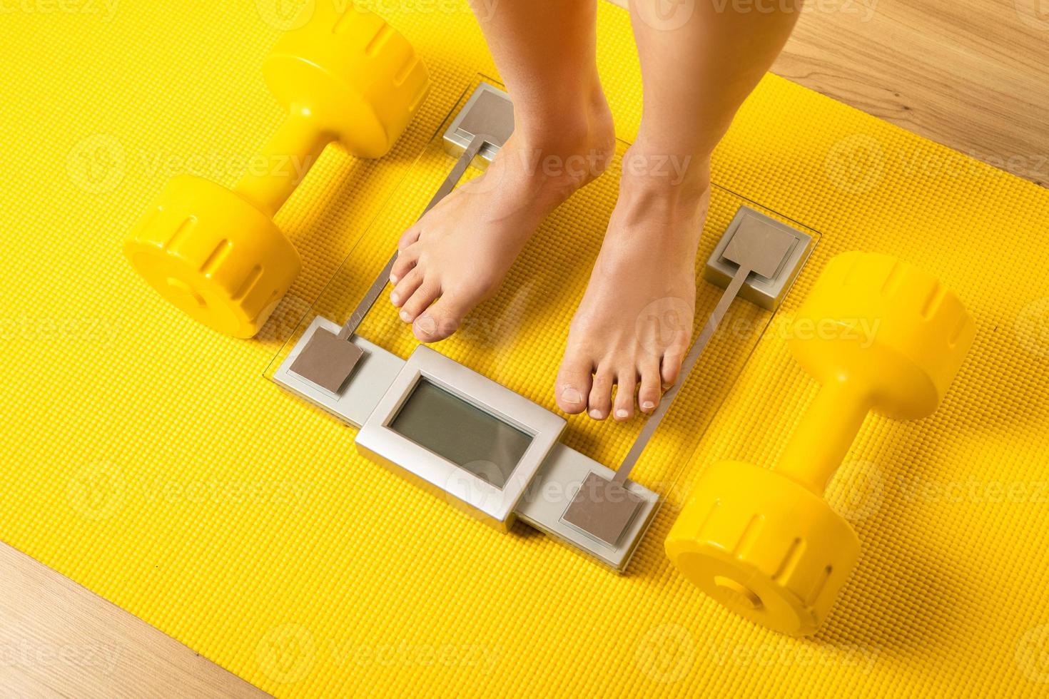 Female feet on the modern weighing scale with a yellow dumbbells and fitness mat photo