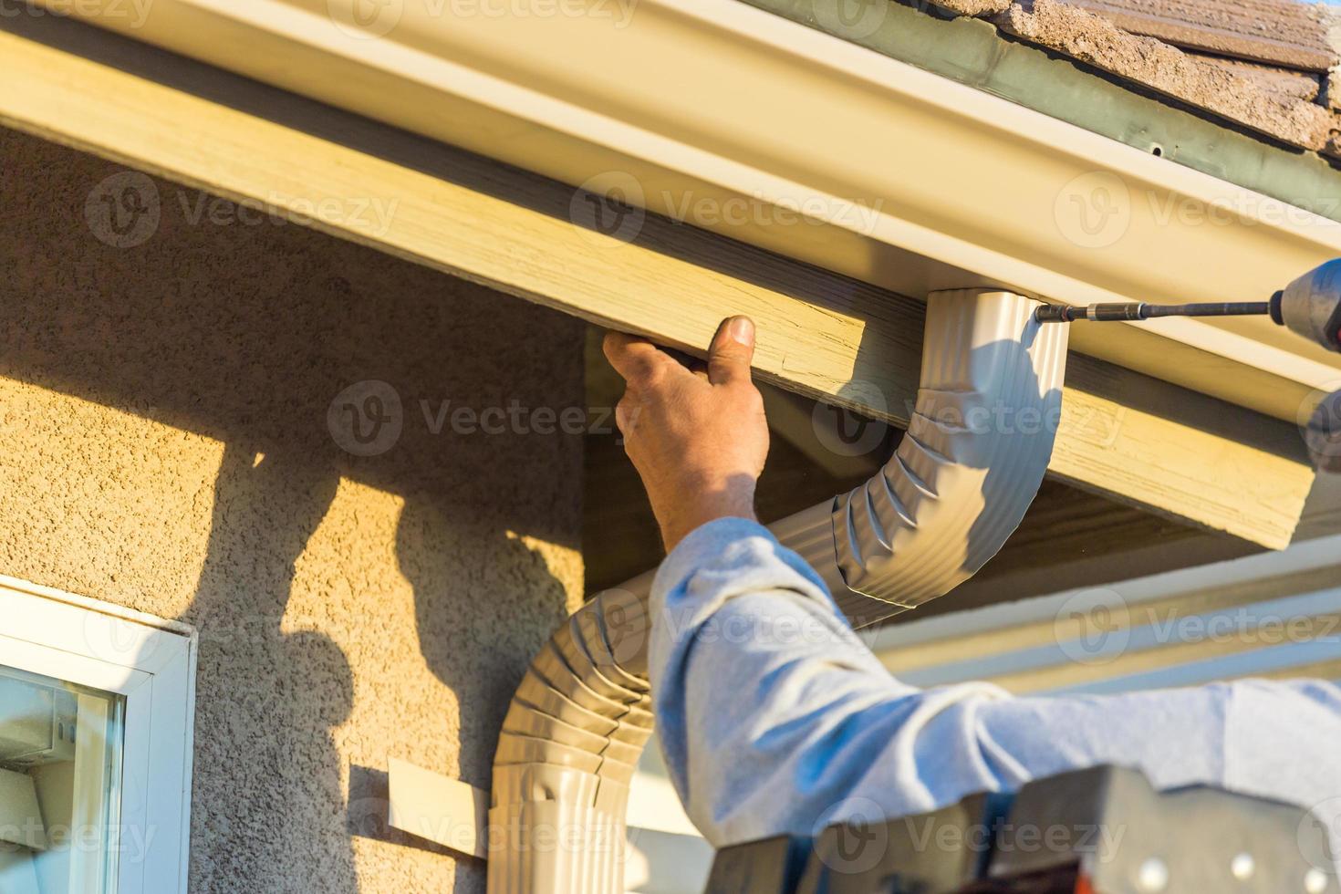 Worker Attaching Aluminum Rain Gutter and Down Spout to Fascia of House. photo