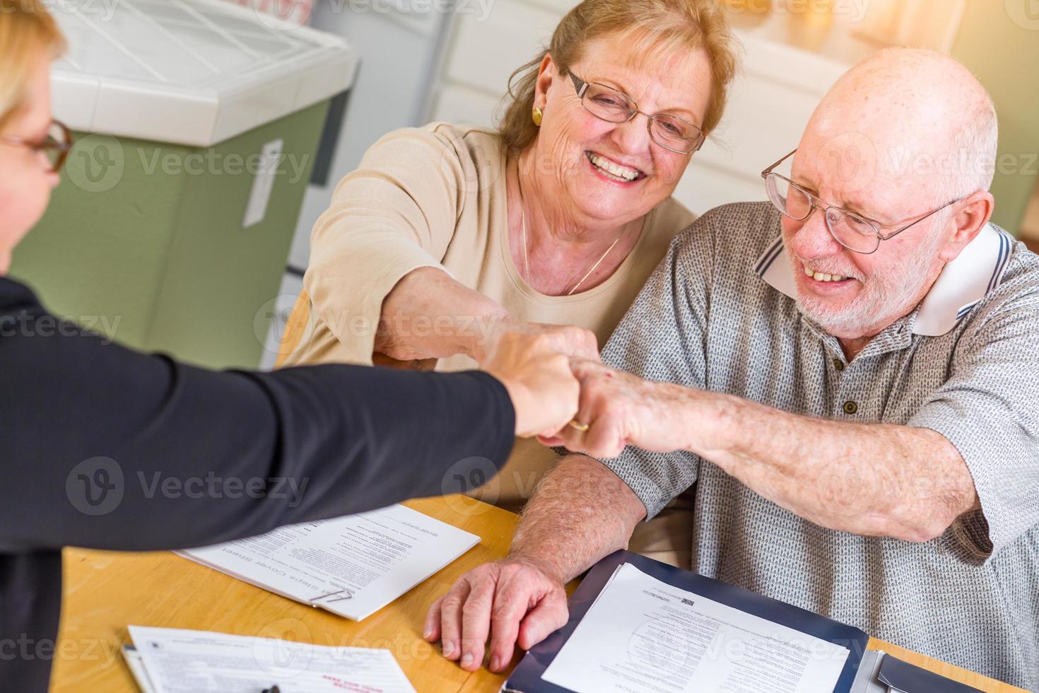 pareja de adultos mayores celebrando con golpes de puño sobre documentos en su casa con un agente al firmar foto