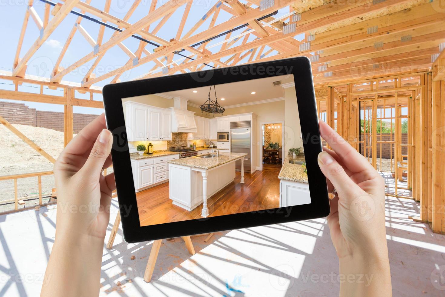 Female Hands Holding Computer Tablet with Finished Kitchen on Screen, Construction Framing Behind. photo