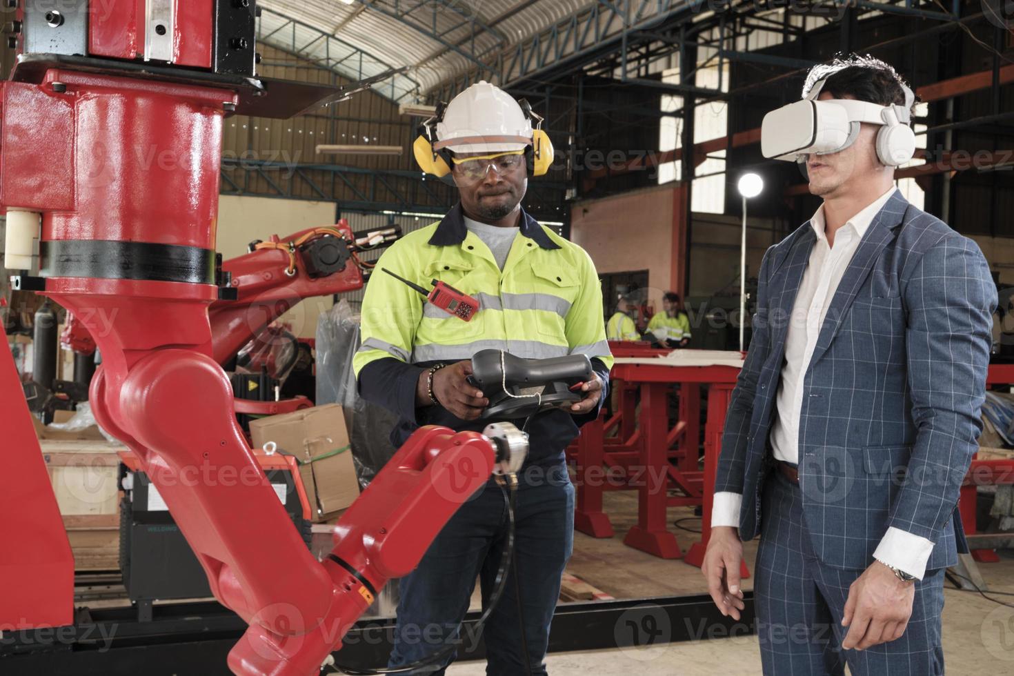 African American engineer demonstrates robotic arm control to customer with VR headsets simulator in mechanical manufacture factory, intelligent industrial machines, and futuristic technology. photo