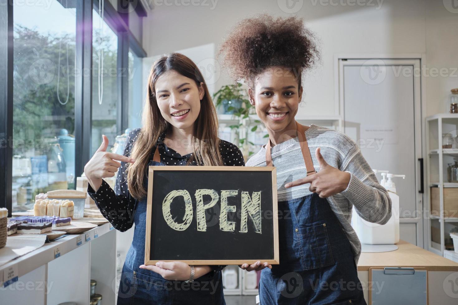 Two young female shopkeepers show open sign board with cheerful smiles in refill store shop, happy work with organic products, zero waste groceries, eco-friendly merchandise, and sustainable business. photo