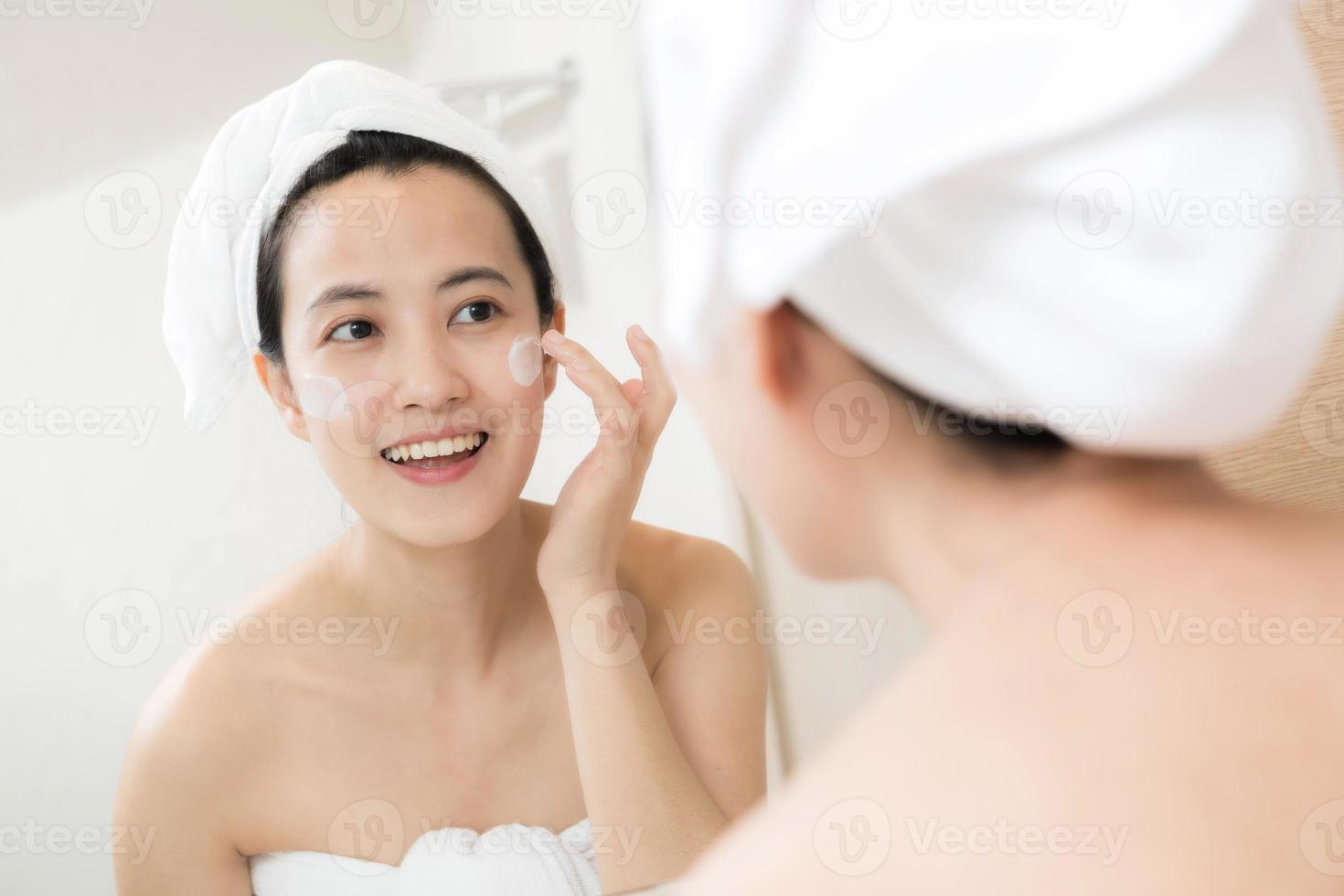 Happy young Asian woman applying face lotions while wearing a towel and touching her face in bathroom photo