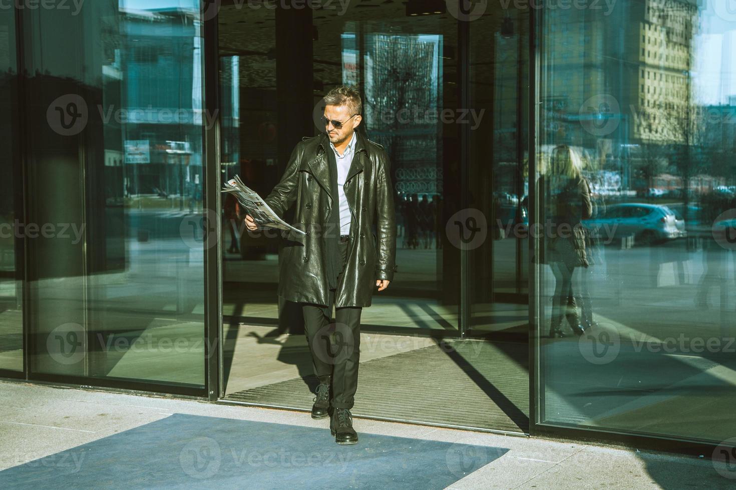 Horizontal photo of beautiful adult male model in sunglasses and leather jacket with newspaper in hand