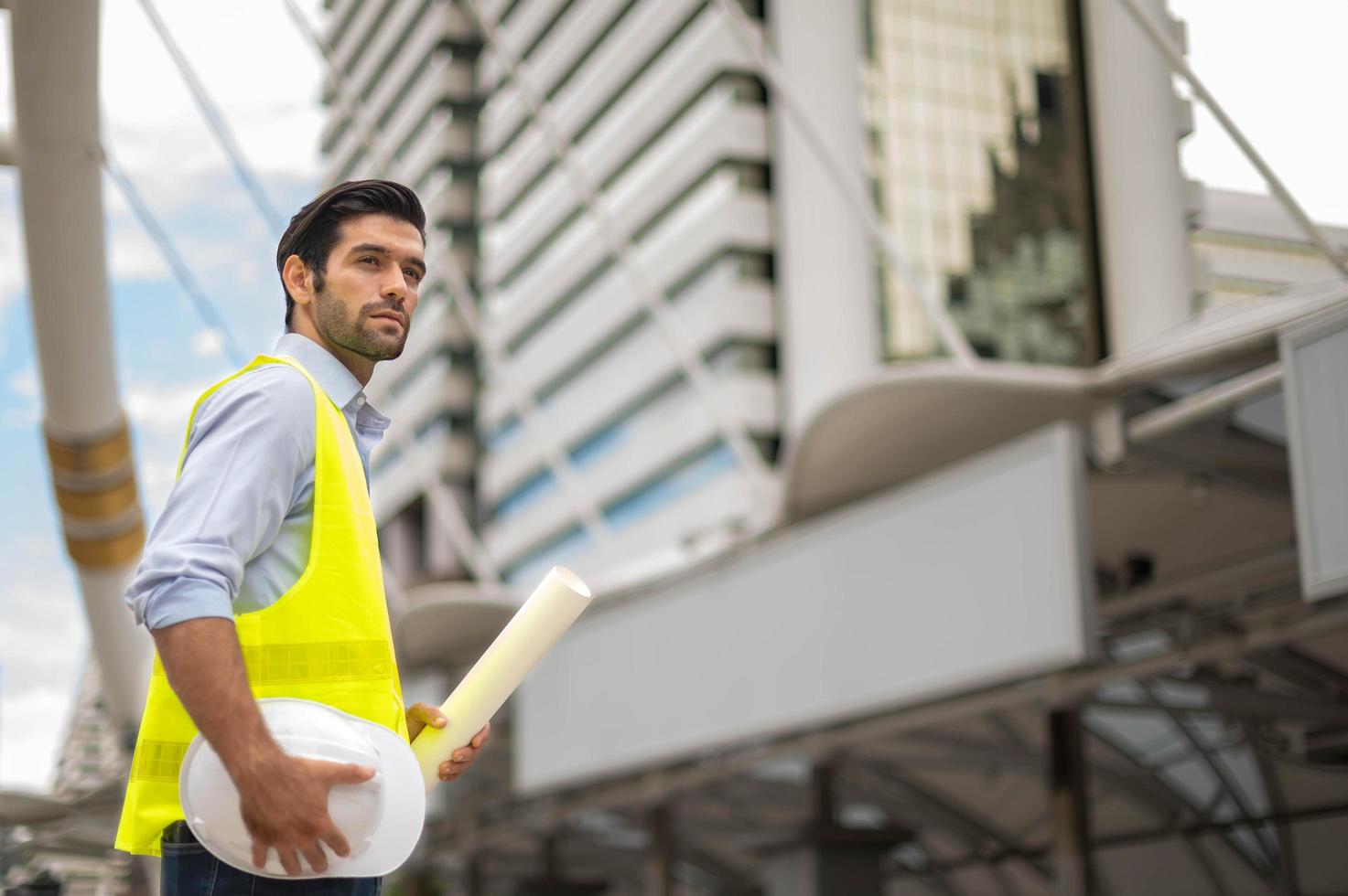 ingeniero caucásico, con chaleco amarillo y sombrero duro grande, y mano sosteniendo el plano de planta blanco en el sitio de trabajo del centro de la ciudad. foto