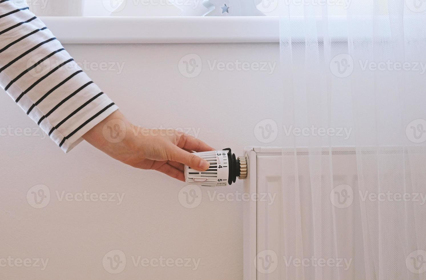 Woman regulates temperature at home with the heating thermostat to save energy, close-up with hand. Temperature control valve on radiator. White heater on wall with curtain in modern apartment. photo