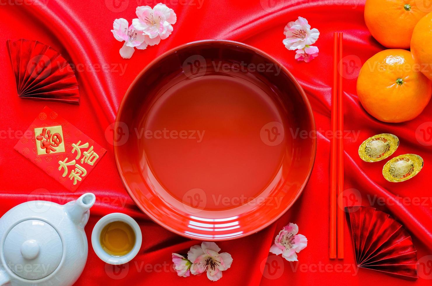 Red plate with chopstick on red satin cloth background with tea set, ingots, oranges and red envelope packet or ang bao word mean weath, lucky for Chinese new year dinner concept. photo