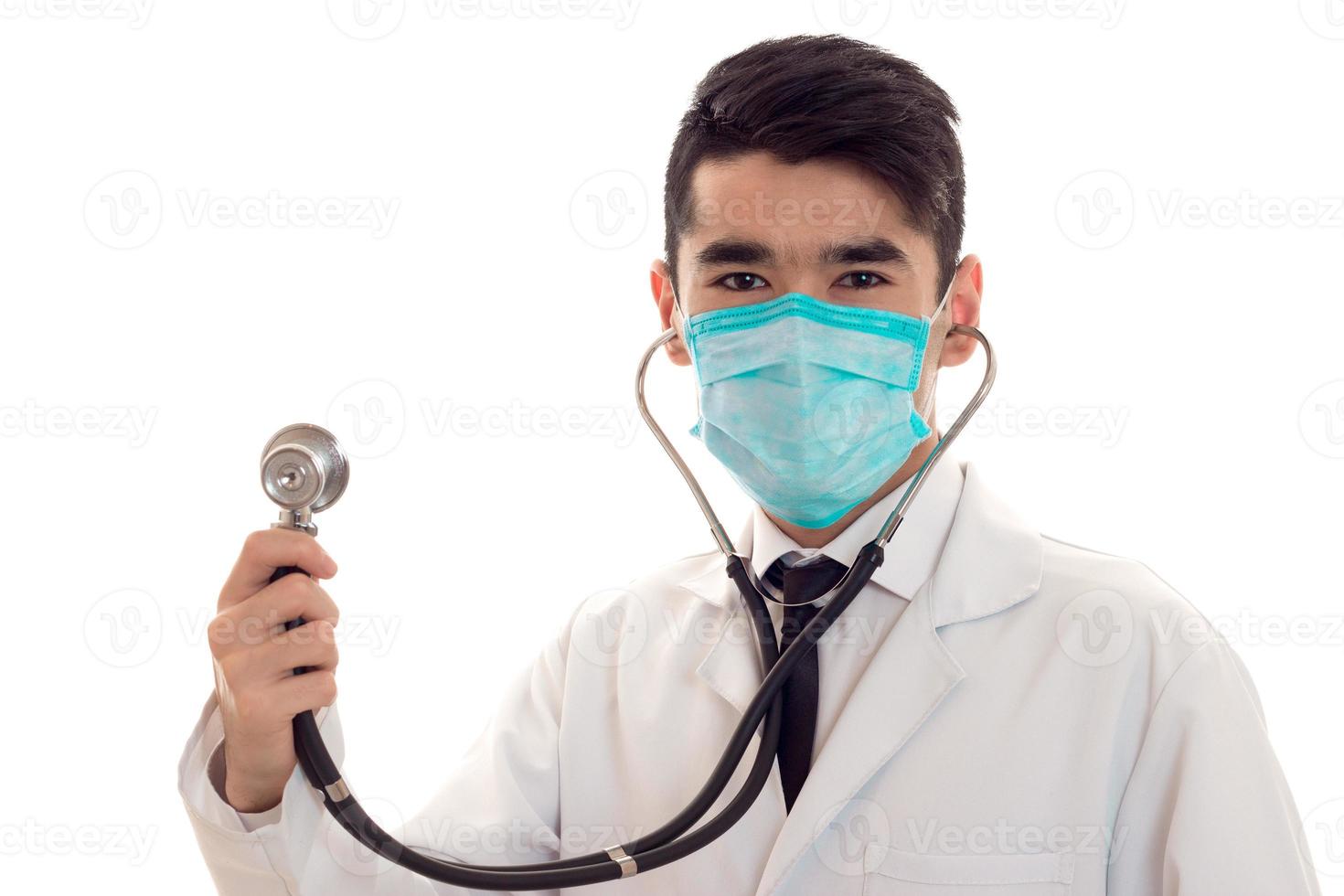 retrato de estudio de médico masculino en uniforme y máscara con estetoscopio en sus manos posando aislado sobre fondo blanco foto