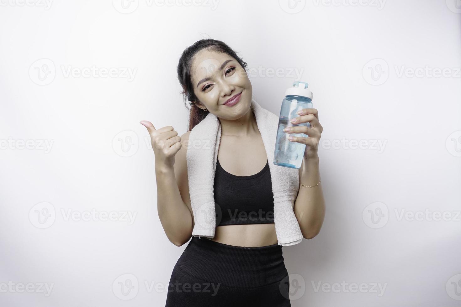 Smiling sportive Asian woman posing with a towel on her shoulder and holding a bottle of water while gesturing OK sign with her finger, relaxing after workout photo