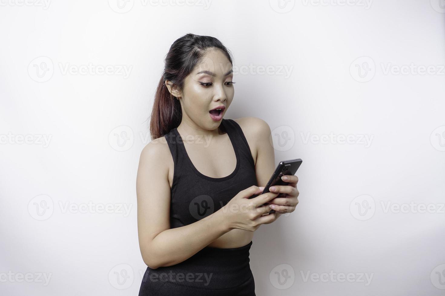 Shocked sporty Asian woman wearing sportswear and holding her phone, isolated by white background photo