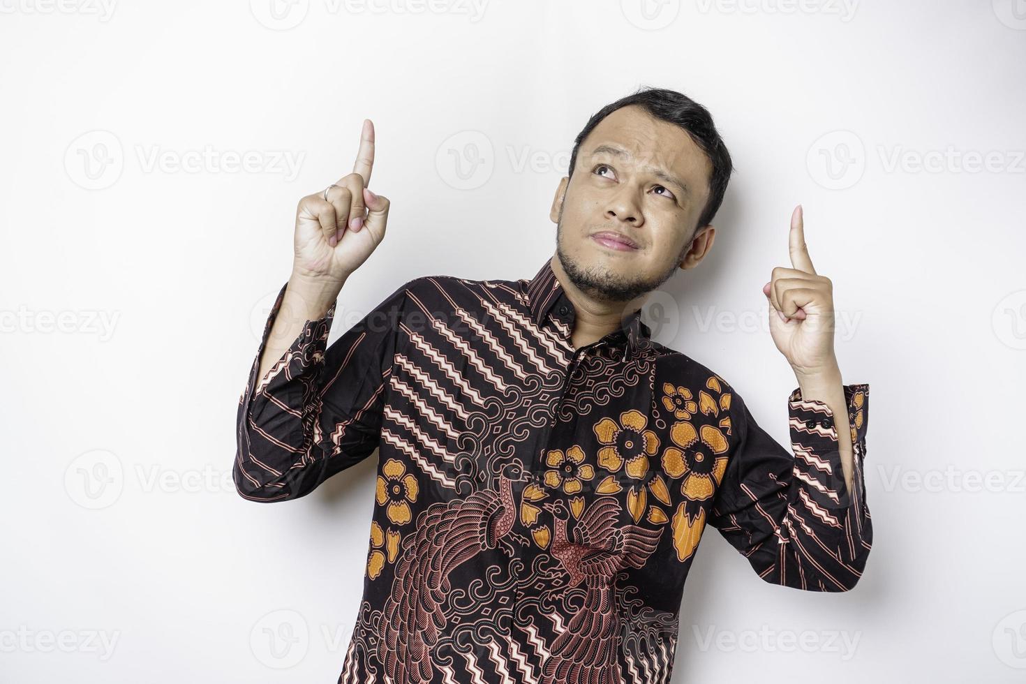 Shocked Asian man wearing batik shirt pointing at the copy space on top of him, isolated by white background photo