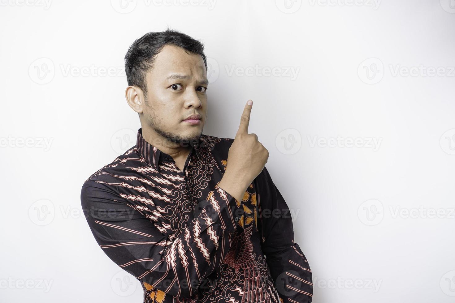 Serious Asian man wearing batik shirt pointing at the copy space on top of him, isolated by white background photo