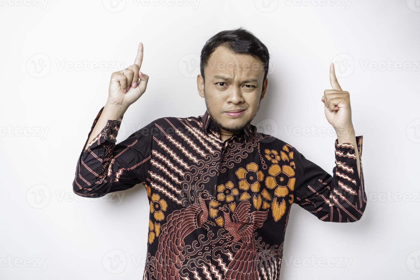 Shocked Asian man wearing batik shirt pointing at the copy space on top of him, isolated by white background photo